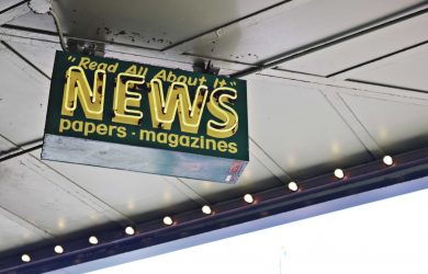 From below of illuminated signboard with news papers magazines inscriptions hanging on metal ceiling on street