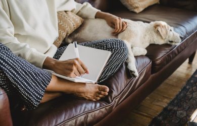 Faceless young female freelancer taking notes while sitting on couch with dog