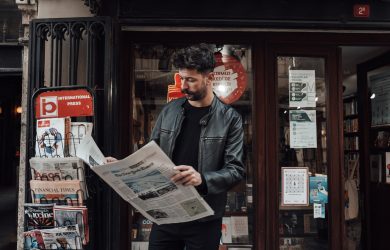 Serious bearded male with trendy haircut in casual clothes reading newspaper while standing near entrance of shop