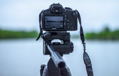 Selective Focus Photo of Black Canon Camera on Tripod Stand in Front of Body of Water Photo