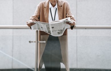 Crop content African American businesswoman in formal clothes reading fresh newspaper and leaning on glass railing of contemporary office building