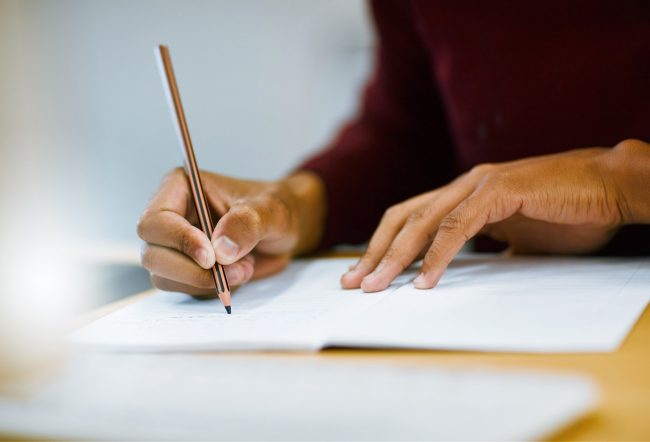 Siedząca osoba przy drewnianym stole, trzymająca ołówek i pisząca na stronie papieru z linią w otwartym notesie