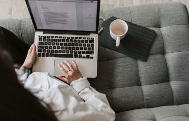 From above of woman in casual wear sitting on comfortable couch with cup and typing on keyboard of laptop while working remotely in cozy living room